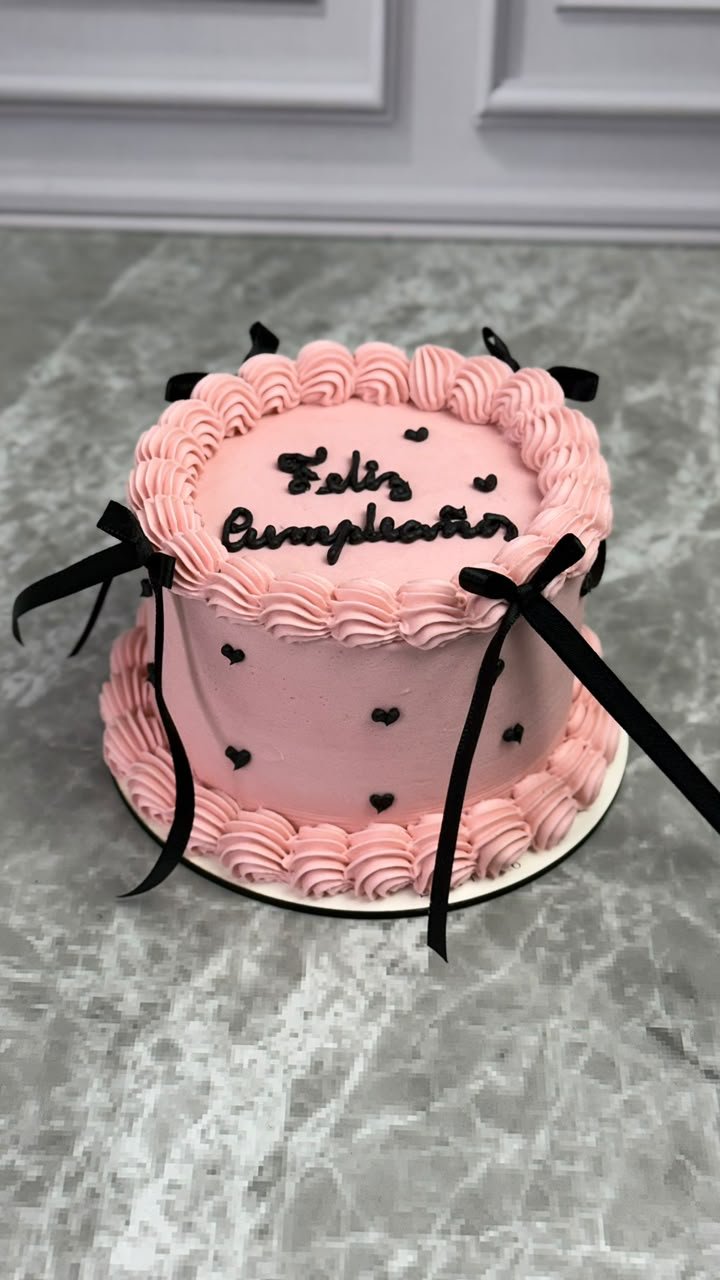 Bolo vintage rosa com bico de confeitar, corações e laços pretos e a frase Feliz cumpleaños