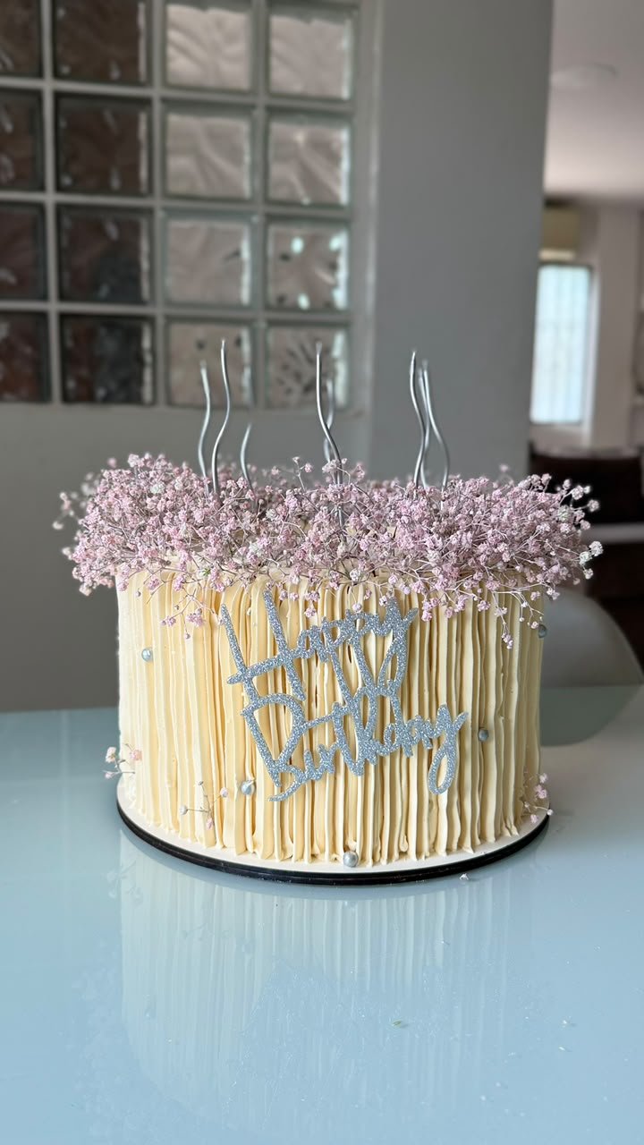 Bolo baixo com cobertura em frisos verticais cor creme, flores mosquitinho rosadas e topo Happy Birthday prateado